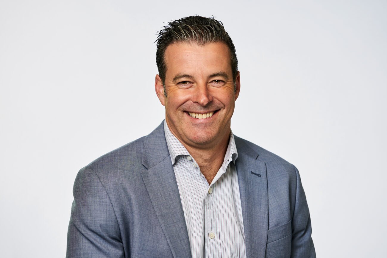 Man smiling at the camera, captured in a corporate headshot, wearing a light gray suit jacket over a white button-up shirt. His hair is short and styled back. The background is plain and light, highlighting his professional demeanor. James Gifford-Mead Photography - Event Photographer London
