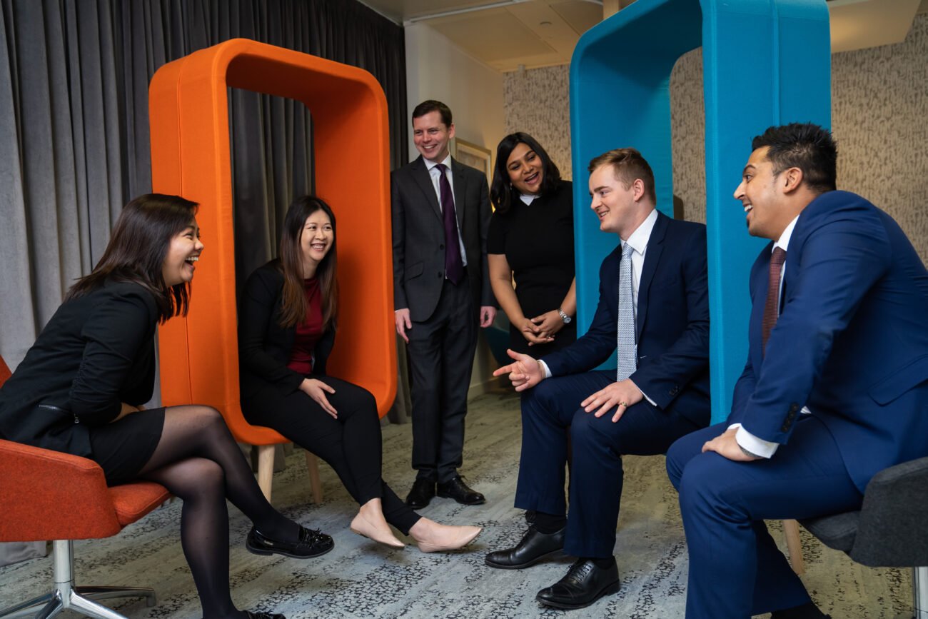 A group of six people in business attire are engaged in a lively discussion captured by a corporate photographer in London. Two women are seated in colorful, phone booth-like chairs. Everyone is smiling and laughing, creating a relaxed, friendly atmosphere in the modern office space