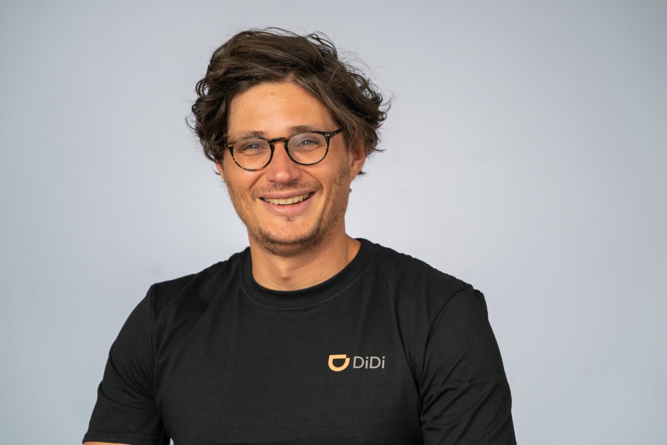 A person with curly hair and glasses smiles at the camera, exuding confidence. They wear a black t-shirt adorned with a "DiDi" logo. Captured with the precision of a corporate headshot photographer, their image stands out against a plain, light gray background. James Gifford-Mead Photography - Event Photographer London