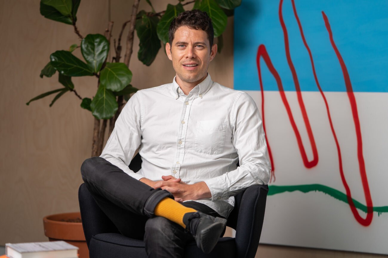 A person in a white shirt and yellow socks sits in a chair, smiling warmly. Captured with the flair of a corporate headshot photographer, the scene includes a large, abstract painting with red and blue elements and a potted plant with broad leaves in the background. James Gifford-Mead Photography - Event Photographer London