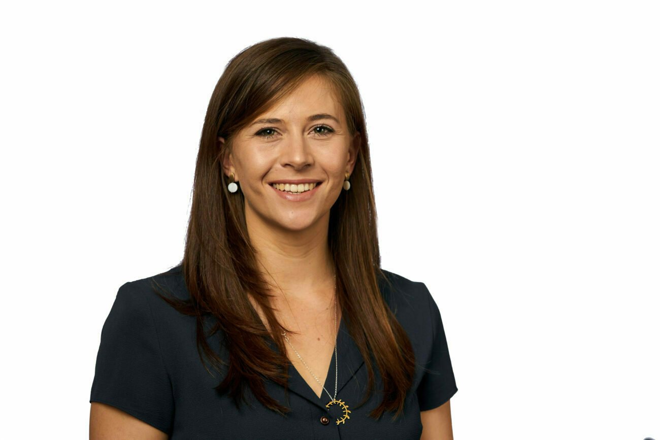 A woman with long brown hair and a navy blouse stands smiling against a white background, captured by a corporate headshot photographer. She sports round earrings and a sun-shaped pendant necklace. James Gifford-Mead Photography - Event Photographer London