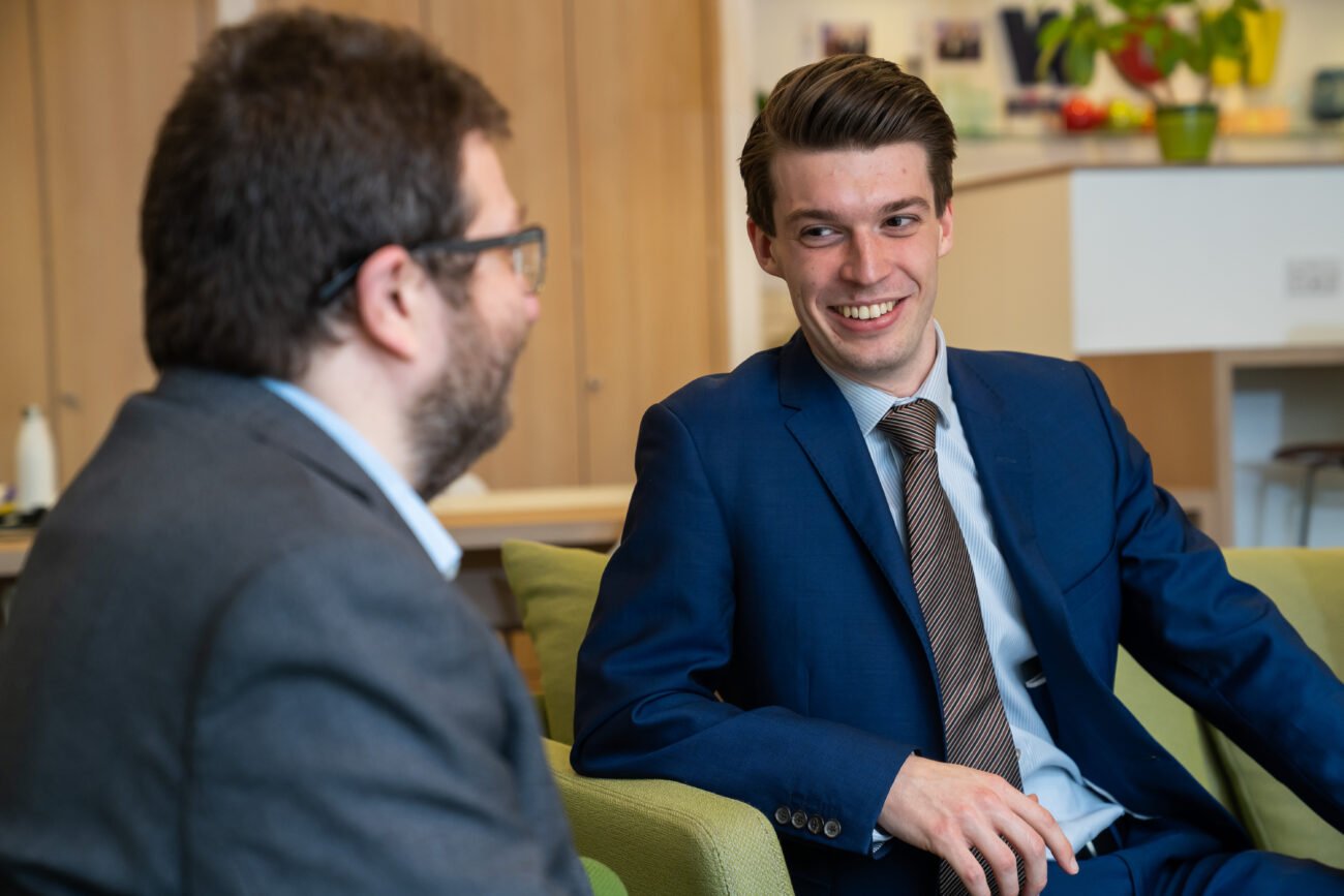 A corporate photographer in London might capture this scene: two men in suits engaging in conversation within an office, the man on the right, clad in a blue suit, smiles at his bespectacled counterpart in a gray suit. Shelves and a countertop adorned with a plant complete the background