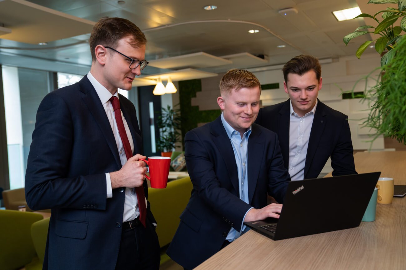 In a modern office setting captured by a corporate photographer in London, three people gather around a laptop. One holds a red mug, another types, as the third gazes at the screen. They appear focused amidst plants and comfortable seating in the background