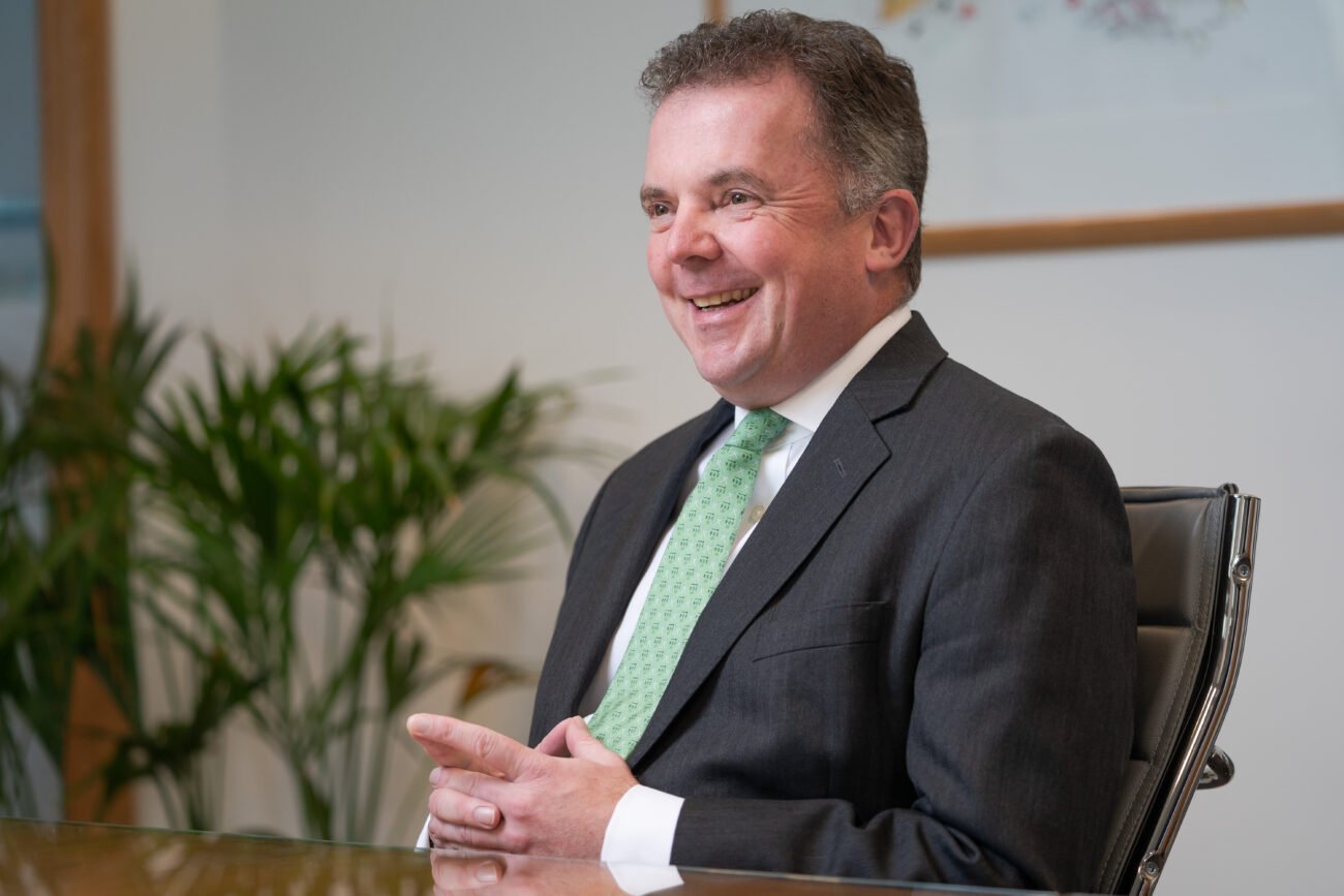 A man in a suit and green tie sits at a table, smiling and gesturing with his hands. Captured by a corporate photographer in London, the scene includes a plant and framed artwork in the background