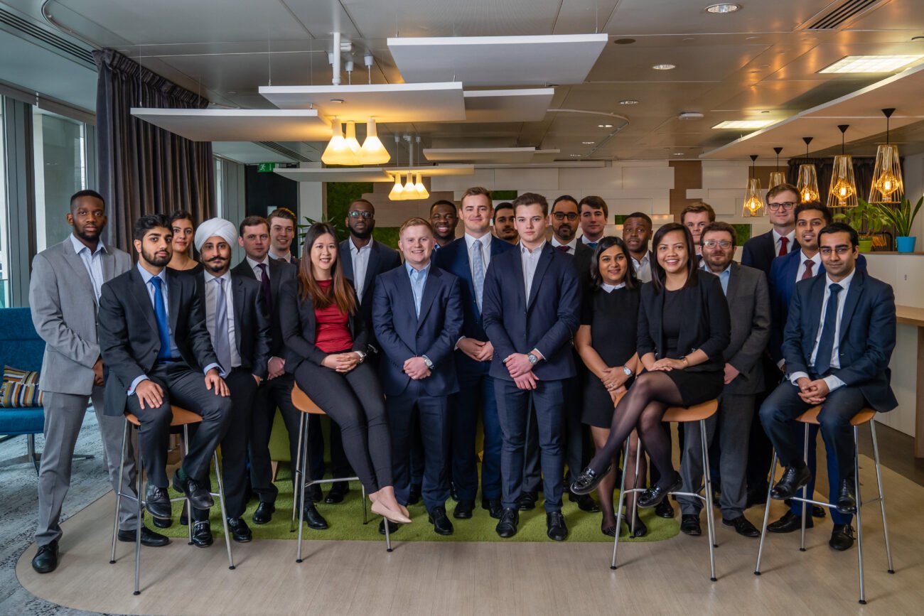 A diverse group of professionals, impeccably dressed, gather in an office setting captured by a corporate photographer from London. They pose confidently on bar stools amid a modern interior accentuated by plants and stylish ceiling lights