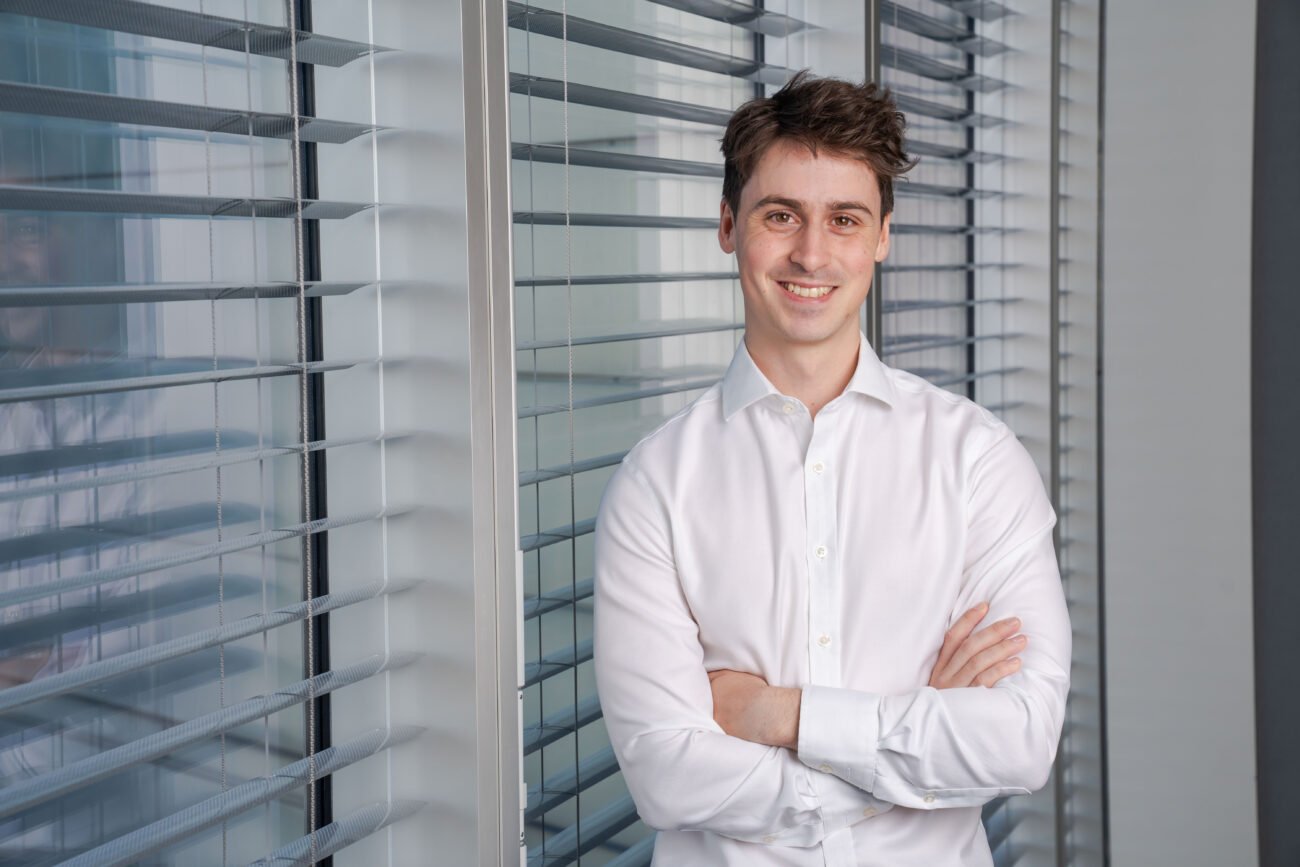 A smiling man in a white shirt stands confidently with arms crossed, bathed in sunlight filtering through the blinds of large windows. Captured by a corporate headshot photographer, he gazes directly at the camera, exuding professionalism and charisma. James Gifford-Mead Photography - Event Photographer London