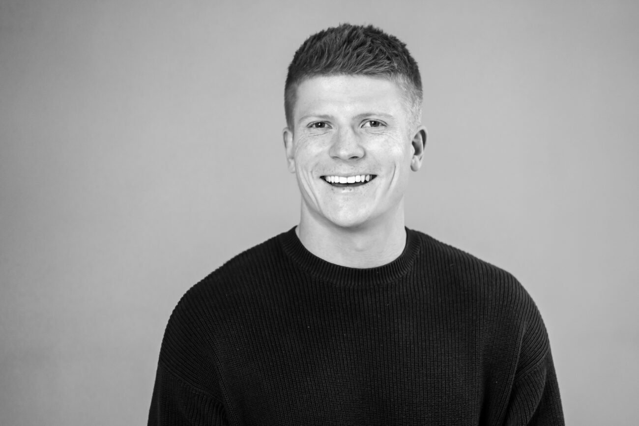 A smiling person with short hair in a black sweater faces the camera, captured against a plain background. This black-and-white image embodies the timeless quality that a skilled corporate headshot photographer can achieve, conveying professionalism and approachability. James Gifford-Mead Photography - Event Photographer London