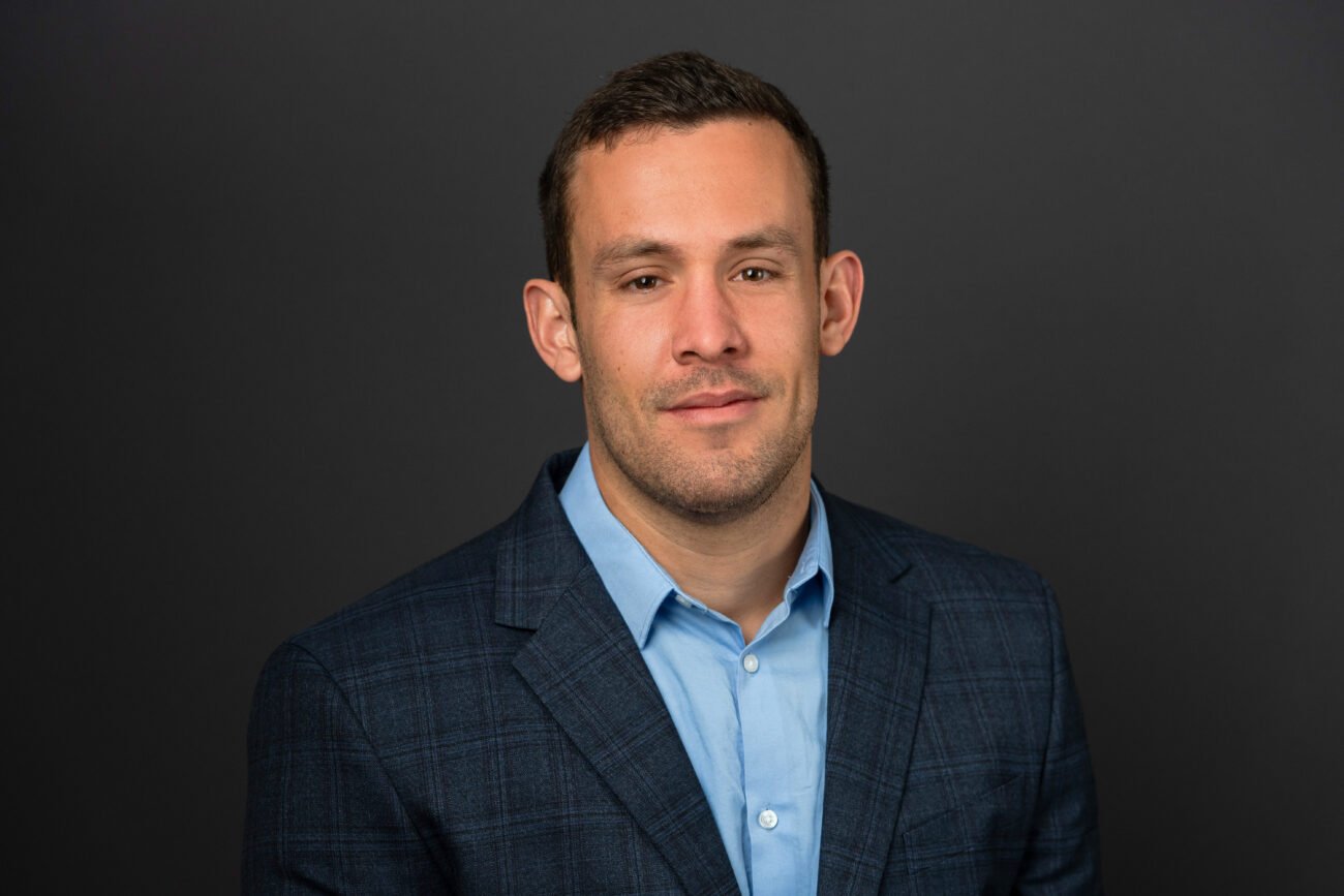 A man in a blue checkered suit and light blue shirt poses against a dark gray background. With short brown hair and a slight smile, he presents a polished image perfect for any corporate headshot photographer's portfolio. James Gifford-Mead Photography - Event Photographer London
