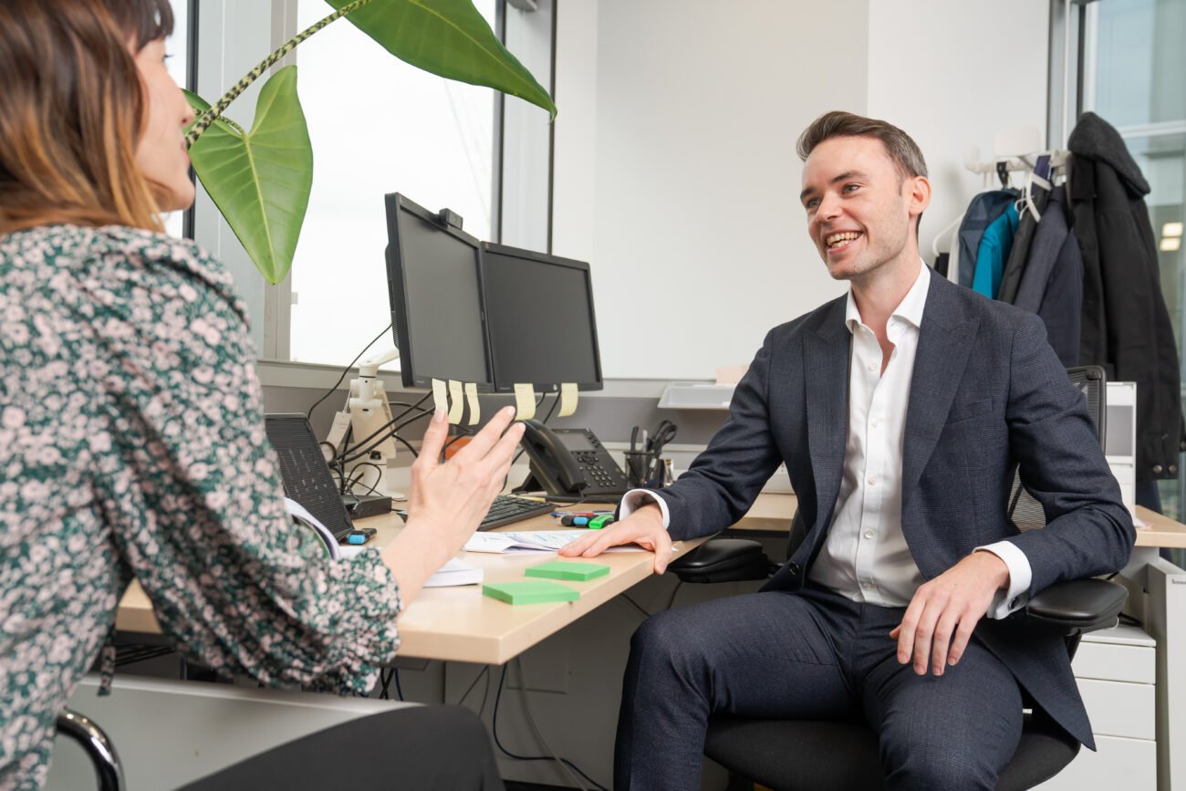 Two professionals engage in a lively discussion, captured by a corporate photographer. The man in a suit smiles at the desk, while the woman in her floral blouse gestures animatedly. Behind them, a large plant and dual computer monitors complete this quintessential London office scene