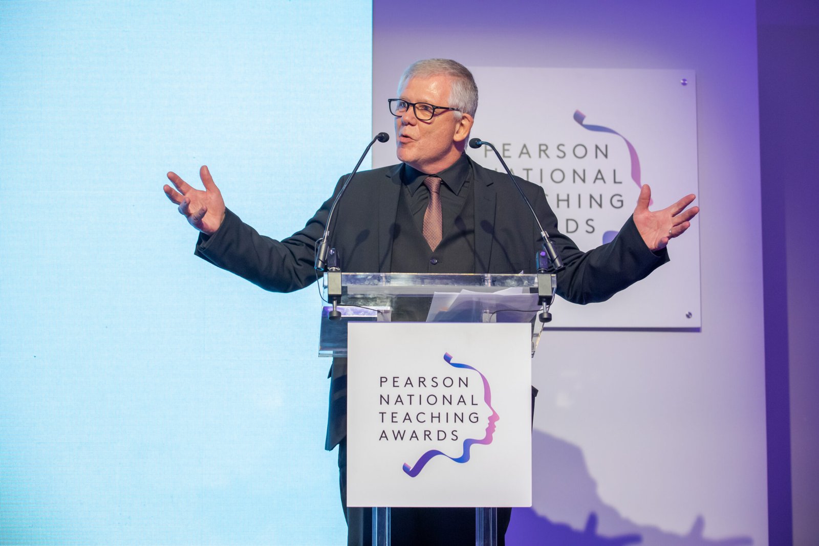 A gray-haired man in glasses speaks passionately at a clear podium, hands outstretched. Behind him are signs for the Pearson National Teaching Awards. He's wearing a dark suit and tie against a blue and purple backdrop. James Gifford-Mead Photography - Event Photographer London
