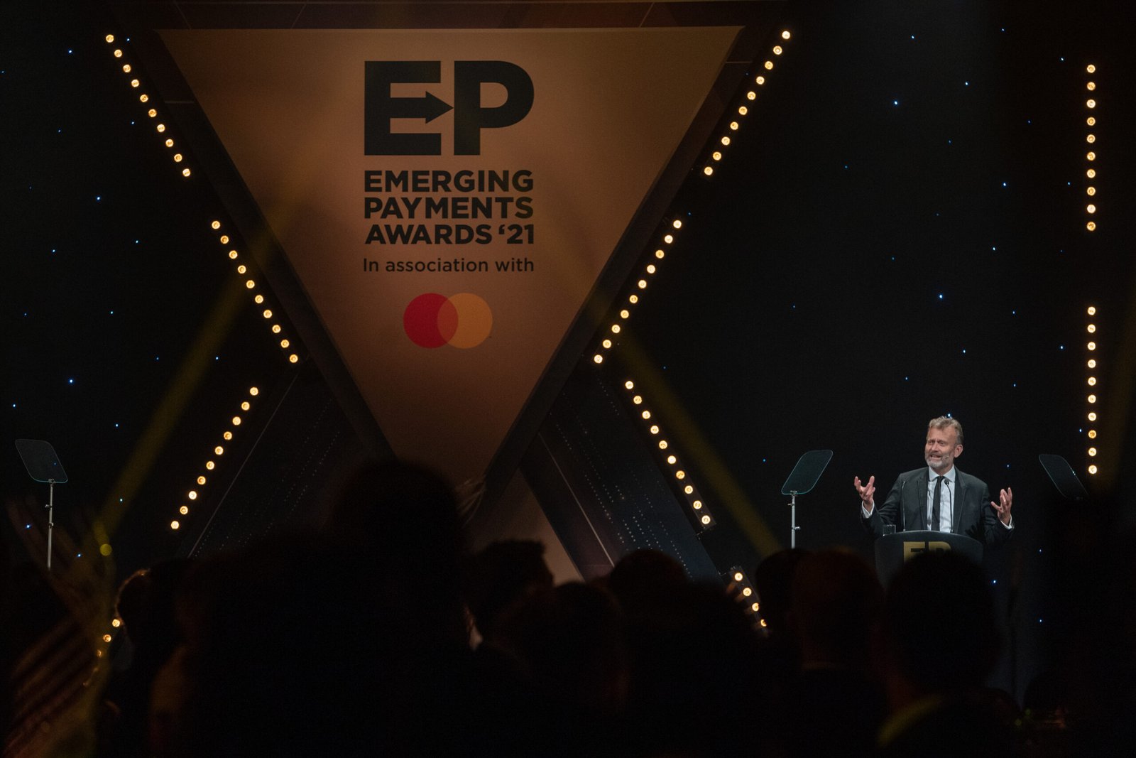 A person speaks at a podium on a dimly lit stage at the Emerging Payments Awards 2021. The background features a large triangular display with 