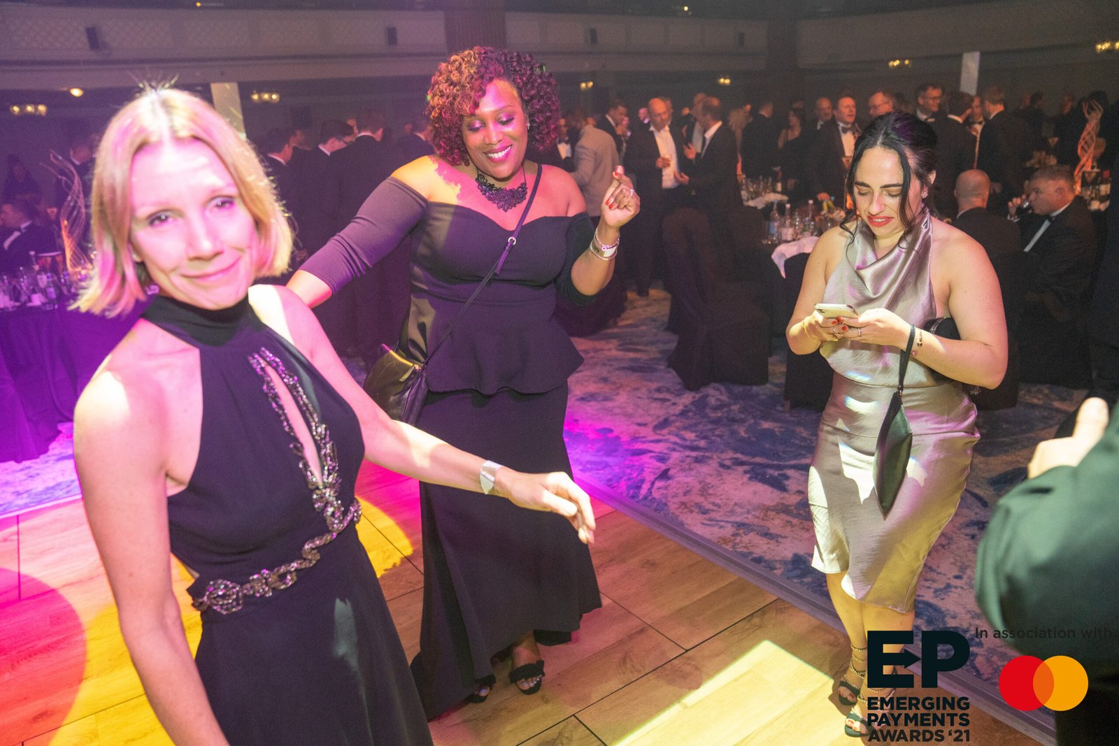 Three women are on a dance floor at a formal event. Two are dancing and smiling, while the third is looking at her phone. People in formal attire are seated in the background. There's event branding with 