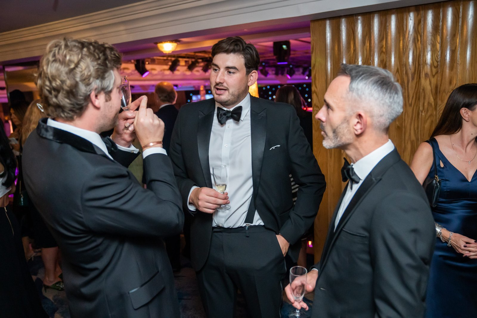Three men in formal attire converse at an indoor event. They are wearing suits and bow ties, holding drinks. The background shows other attendees and purple lighting, suggesting a social or formal gathering. James Gifford-Mead Photography - Event Photographer London