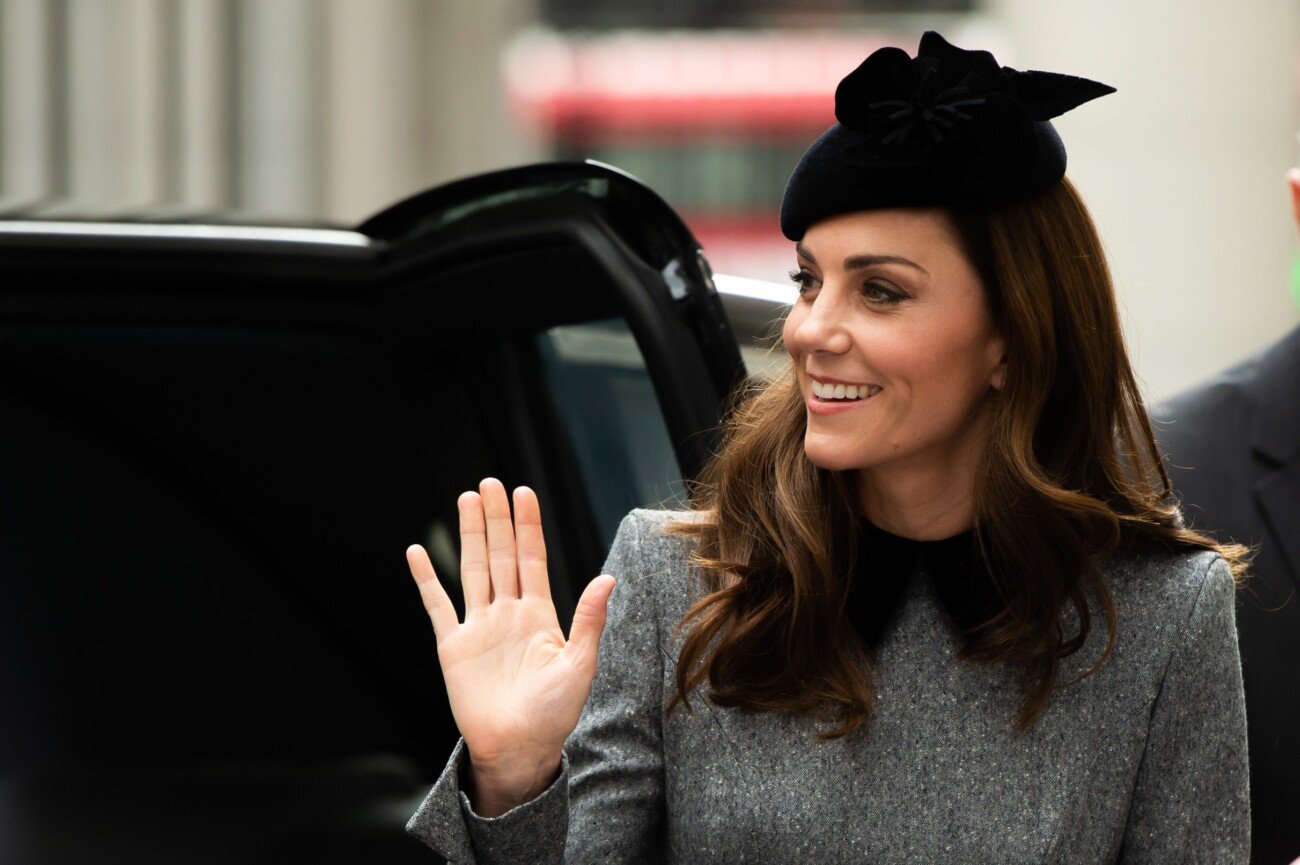 A woman wearing a black hat and gray coat waves outside a black vehicle. Her long brown hair is loose, and she is smiling warmly. The background is slightly blurred, with a glimpse of a red and white object