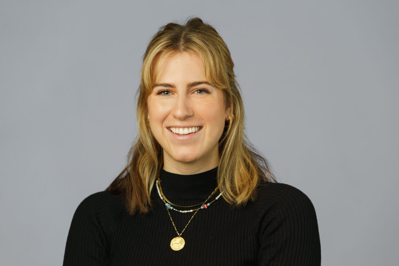 Person with shoulder-length hair smiles confidently at the camera, embodying the refined touch of a corporate headshot photographer. They are wearing a black sweater and layered necklaces against a plain gray background.