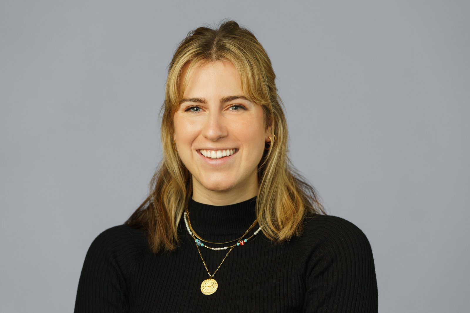 A smiling person with shoulder-length blonde hair, wearing a black turtleneck and layered necklaces, poses for a corporate headshot against a plain gray background