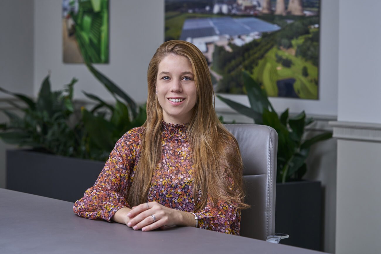 A woman with long hair, wearing a floral blouse, sits at a gray conference table. She smiles and has her hands folded. There are green plants and framed aerial photos on the wall behind her. James Gifford-Mead Photography - Event Photographer London