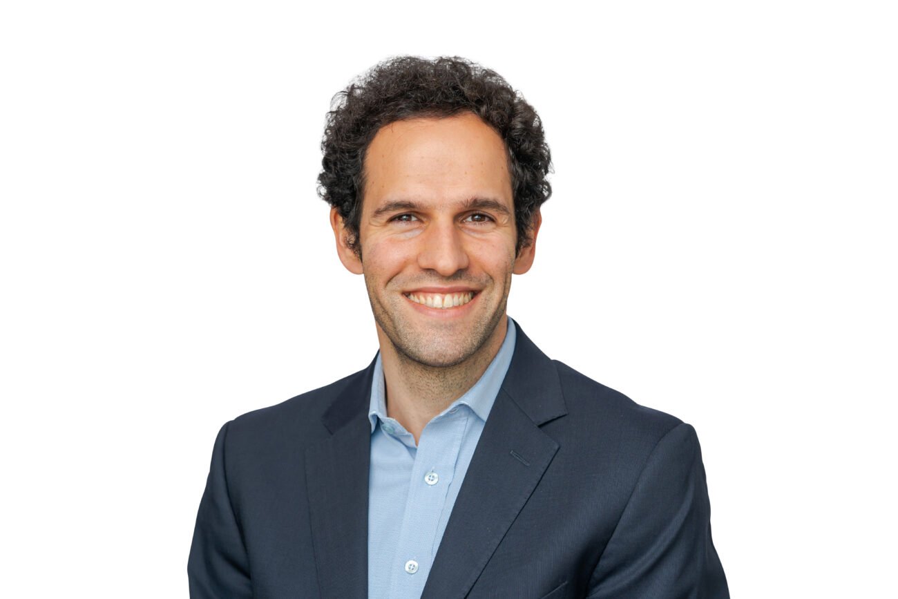 A person with curly dark hair is smiling, perfectly framed by a corporate headshot photographer, wearing a dark suit jacket and light blue shirt. The backdrop is a simple white, emphasizing their professional demeanor. James Gifford-Mead Photography - Event Photographer London