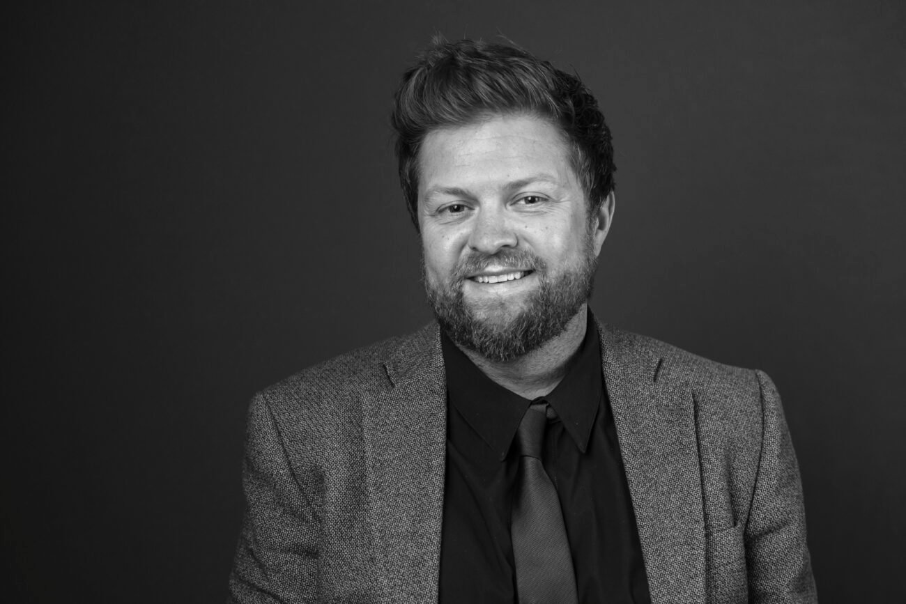 Black and white portrait captured by a corporate headshot photographer: a person with a beard and short hair, wearing a textured blazer over a dark shirt and tie. They are smiling against a plain, dark background. James Gifford-Mead Photography - Event Photographer London
