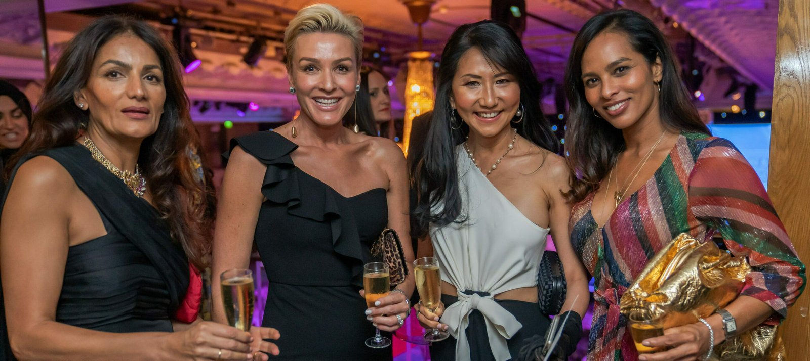 Four women dressed elegantly, holding champagne glasses at an indoor event. The setting is warmly lit with a festive atmosphere. The women are smiling and enjoying the occasion together