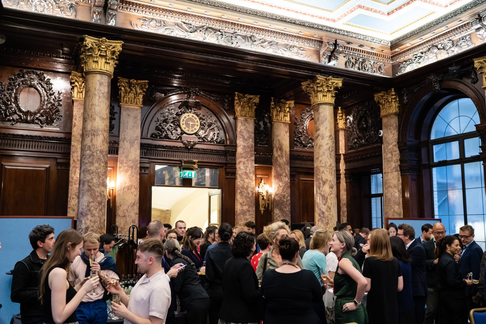A large group of people gather in an ornate room with marble columns and intricate wooden carvings. They are engaged in conversation, holding drinks. The room features high ceilings and elaborate decorations. James Gifford-Mead Photography - Event Photographer London