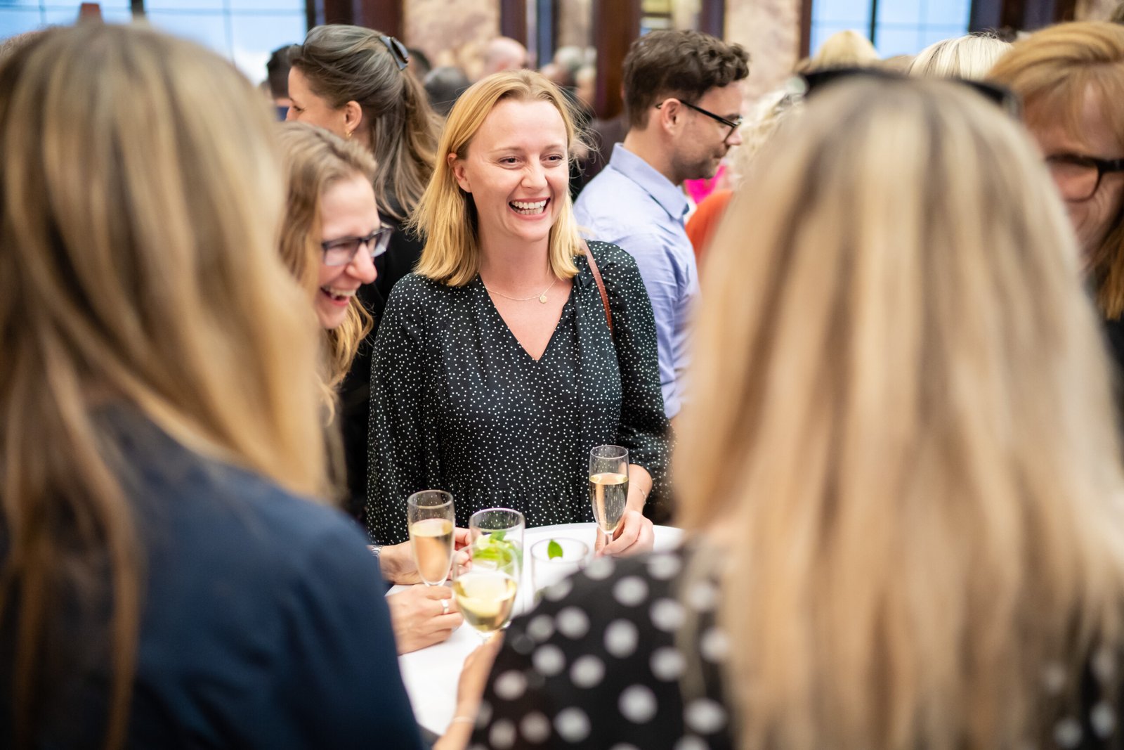 People gathered at a social event, chatting and smiling while holding glasses of champagne. The focus is on a person in a black blouse, joyfully engaging with others. The atmosphere is lively and cheerful. James Gifford-Mead Photography - Event Photographer London