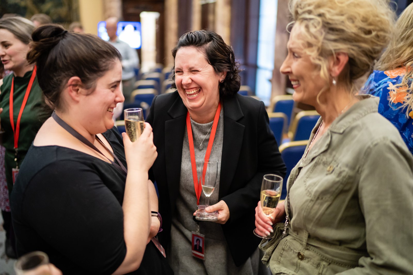 Three women are standing and laughing together, each holding a glass of champagne. They are in a room with other people and empty blue chairs in the background. One woman is wearing a black jacket, another a black top, and the third a green jacket