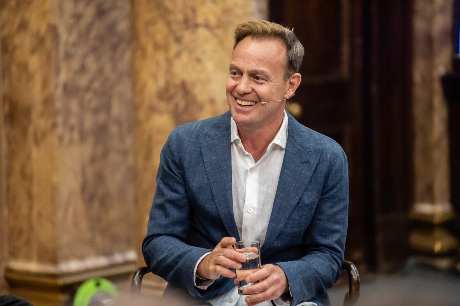 A man in a blue suit and white shirt is sitting and smiling, holding a glass of water. He is wearing a microphone headset, and the background features ornate, marble-like walls