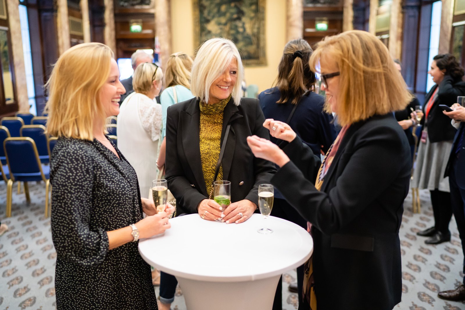 Three women standing and chatting around a tall white table, each holding a glass of champagne. The setting is an elegant indoor venue with ornate decor and several other people in the background. James Gifford-Mead Photography - Event Photographer London