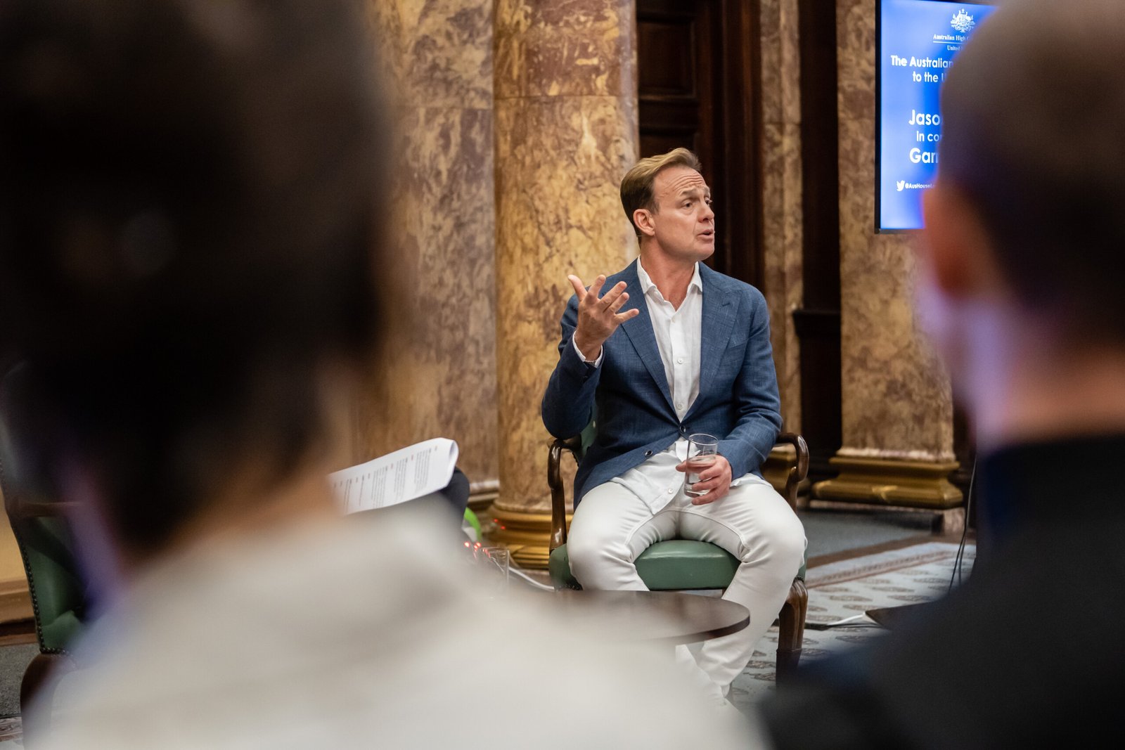 A man in a blue blazer and white pants sits on a stool, gesturing with his hand while speaking. He holds a glass in his other hand. The room has marble walls, and an audience is blurred in the foreground
