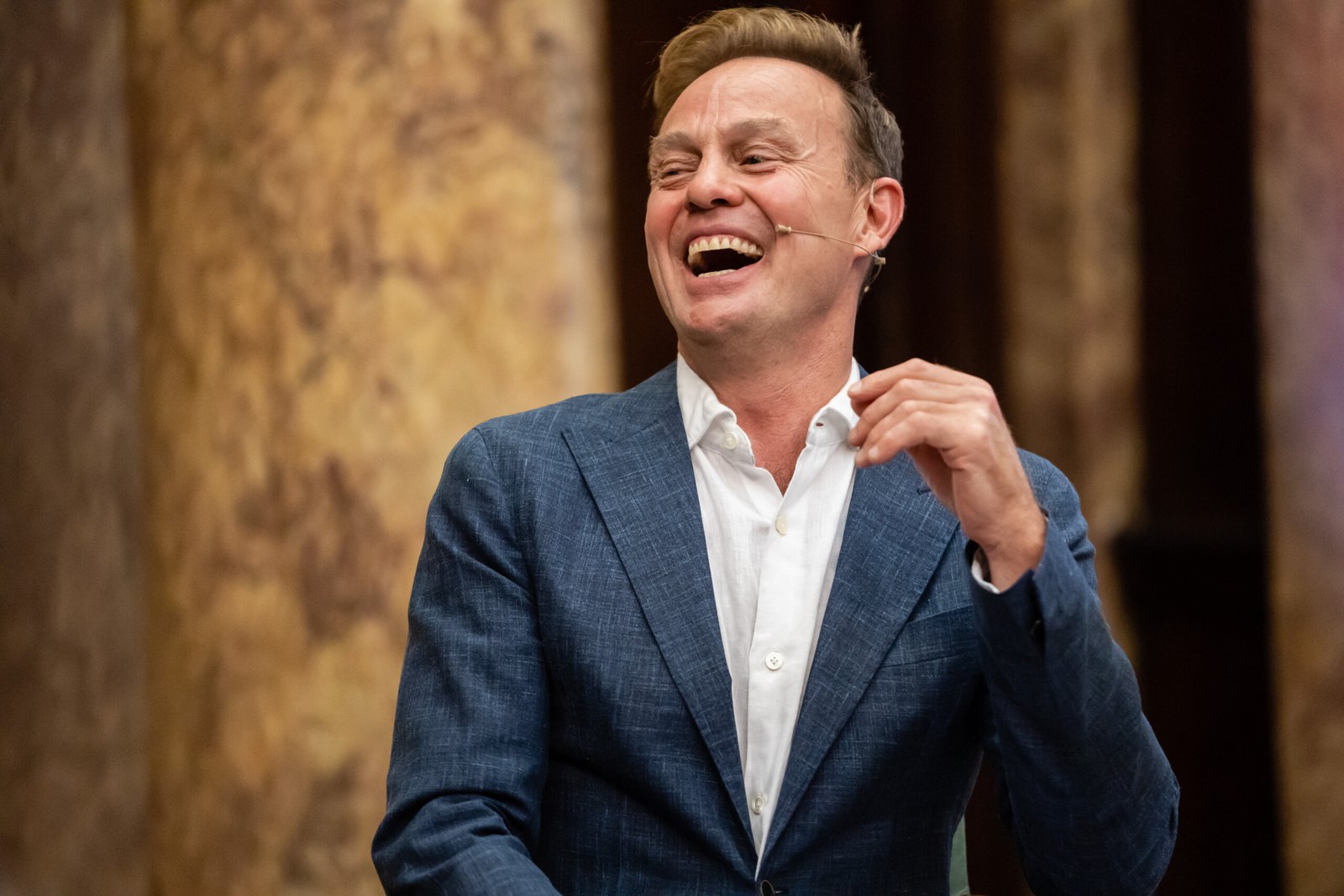 A man in a blue suit and white shirt is laughing while speaking into a headset microphone. He is in front of a blurred, marble-like background with warm tones