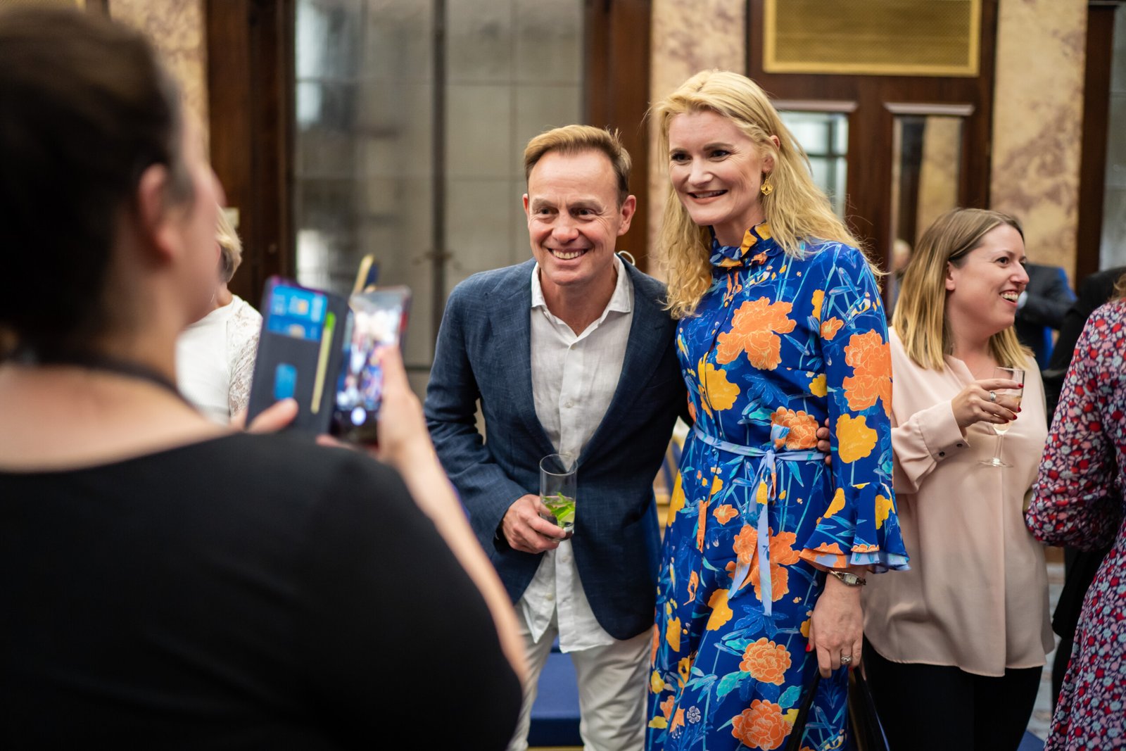 A man and a woman smile for a photo at an indoor event. The woman wears a blue floral dress, while the man wears a blue jacket and holds a drink. Other people in the background are socializing. Another person is taking the photo with a smartphone
