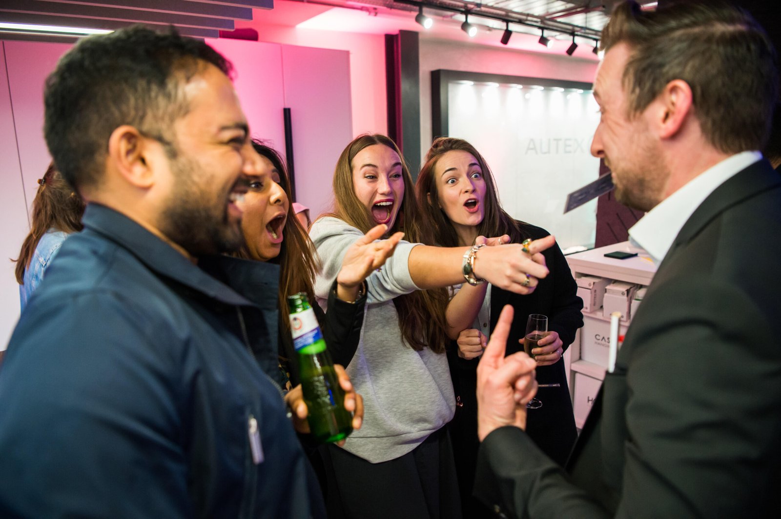 A group of people, including three women and two men, are excitedly pointing and smiling at something out of view. One man holds a beverage, and another holds a drink, engaging in conversation. The setting appears to be indoors at an event