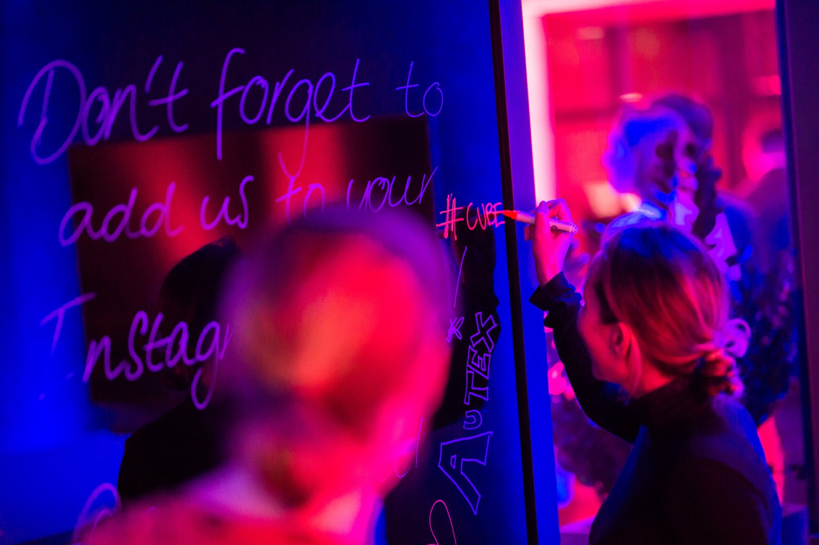 A person in a dimly lit room writes with chalk on a board that says "Don't forget to add us to your Instagram." The room is illuminated with blue and red neon lights