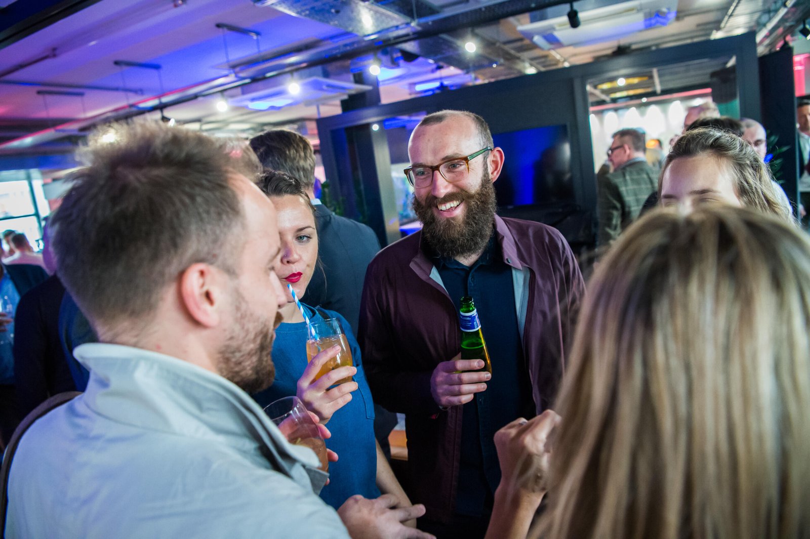 A group of people smiling and conversing at a social event. One man holds a beer bottle, while a woman sips a drink through a straw. The room is brightly lit with colorful lights, creating a lively atmosphere