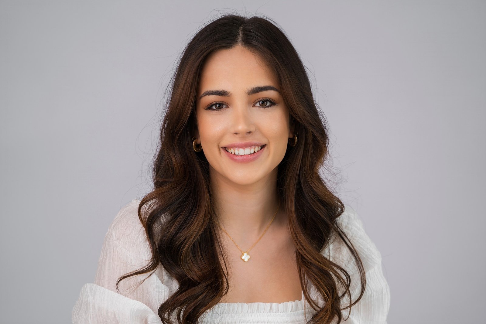 Smiling woman with long, wavy dark hair. She wears a white blouse and a gold necklace with a small pendant. The background is a light gray, providing a clean and simple backdrop