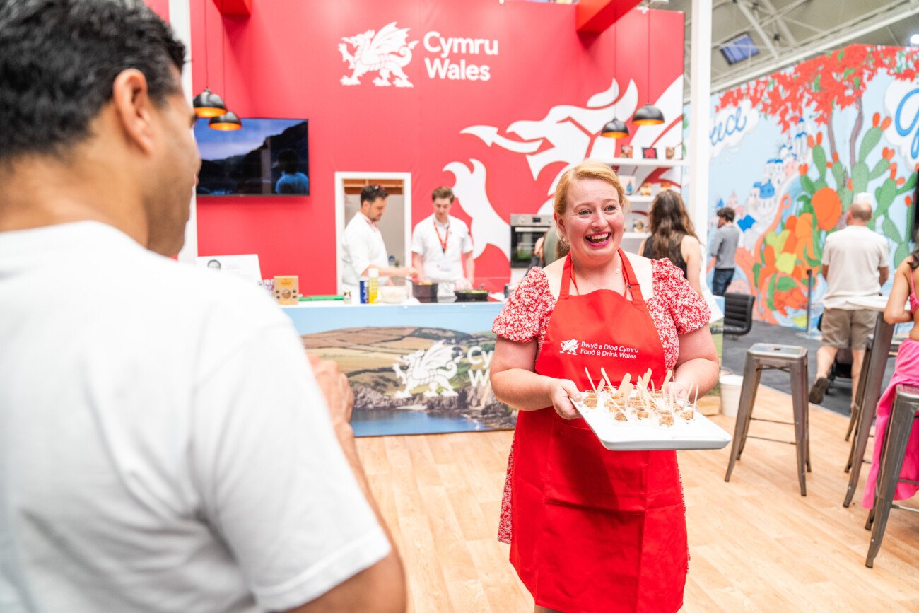 A woman in a red apron smiles while holding a tray of food samples in front of a red booth with "Cymru Wales" written on it. A man stands nearby, with others in the background. The setting is a lively exhibition or fair