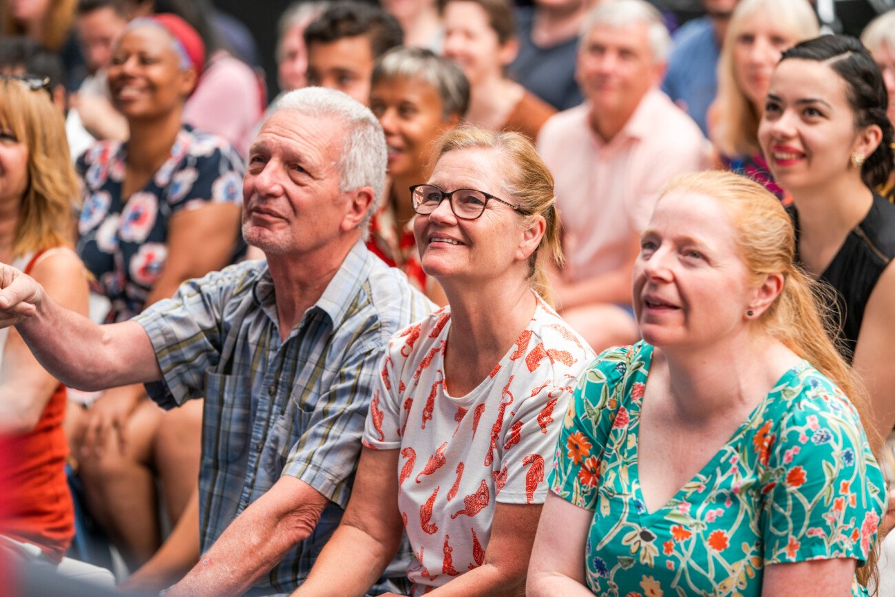 A diverse group of people seated closely together, looking forward and smiling. The focus is on a few individuals in the front row, featuring men and women of various ages and ethnicities, creating a lively and engaging atmosphere