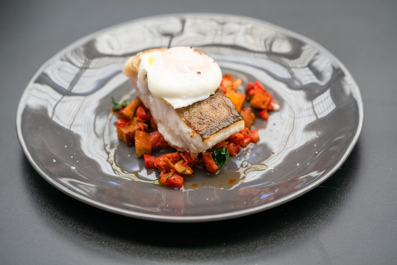 A gourmet plate featuring pan-seared fish with crispy skin, topped with a poached egg. It rests on a colorful bed of diced vegetables, including red peppers. The plate is elegant with reflections on the black glossy surface