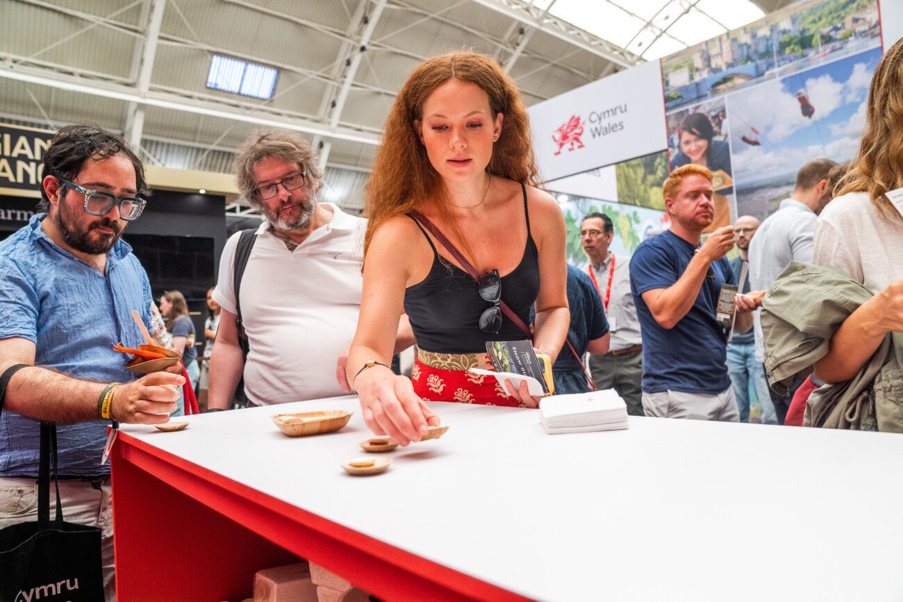 A woman is placing a small item on a white counter at a busy indoor event. She holds a booklet in her other hand. Several people in casual attire are gathered around, engaging and observing. There's a sign with "Cymru Wales" in the background