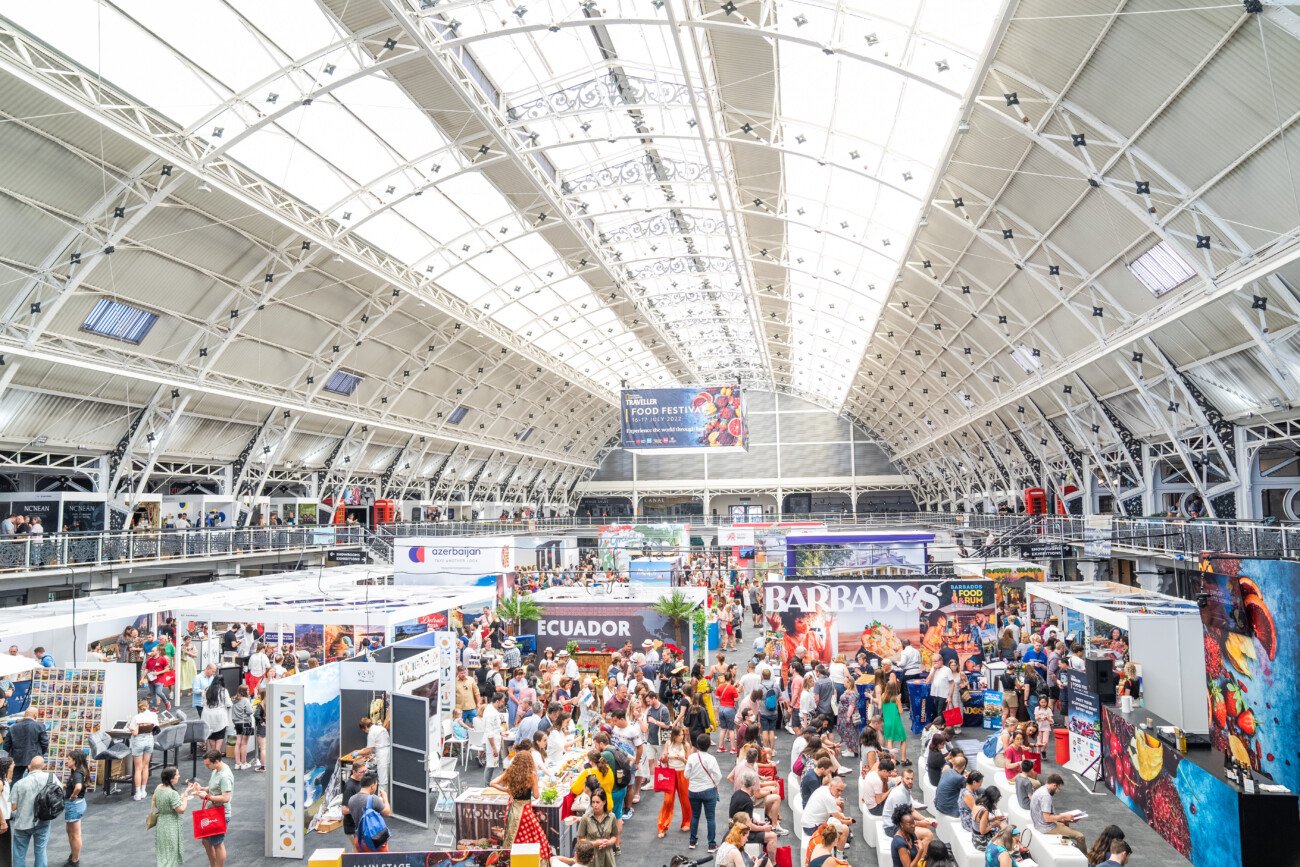 A bustling indoor convention with numerous attendees exploring various booths from different countries, such as Ecuador and Barbados. The venue features high ceilings and an arched glass roof, creating a bright and open atmosphere