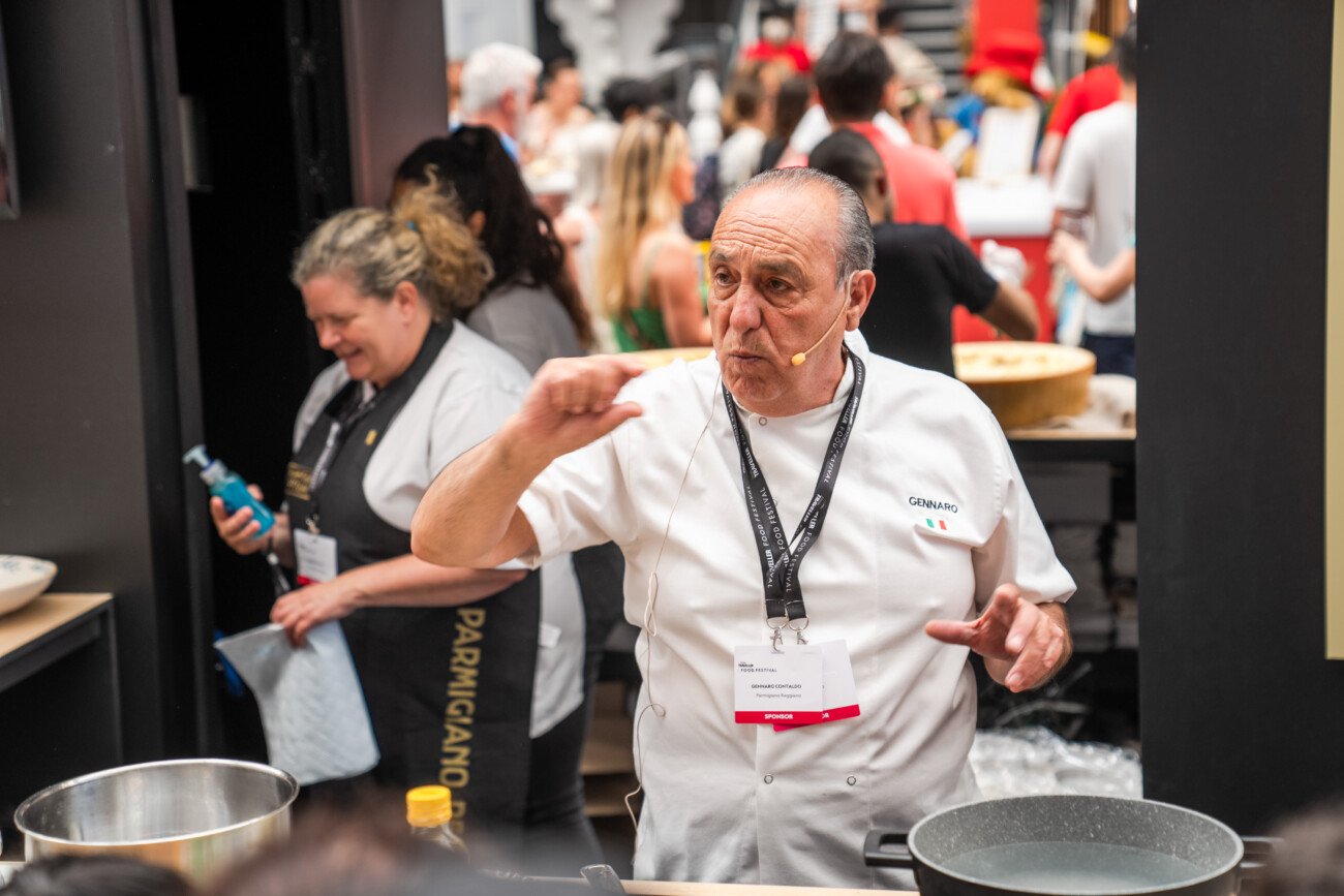 A chef wearing a white uniform and microphone gestures while speaking at a cooking event. Behind him, a woman in a black apron and several attendees are visible. Cooking equipment and ingredients are on the counter in front of the chef