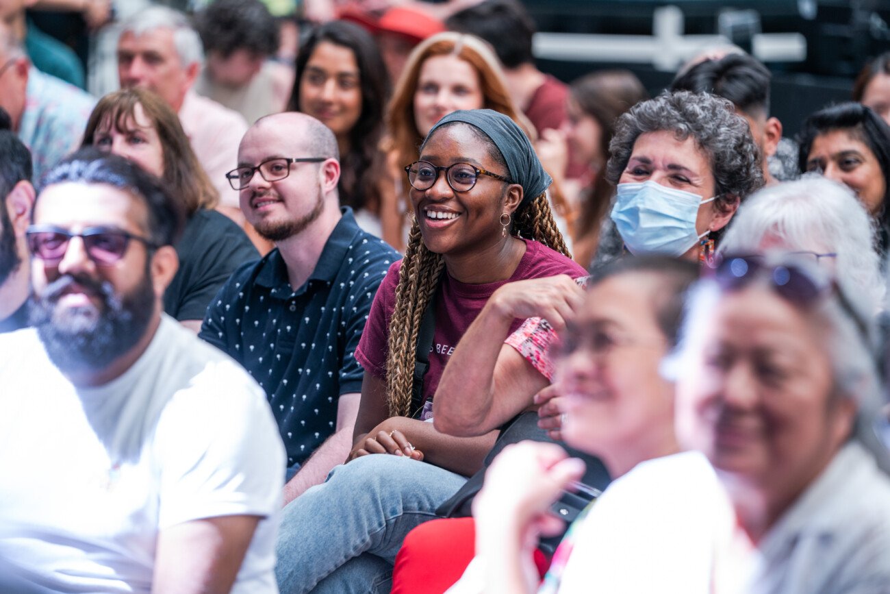 A diverse group of people sit closely together in an audience, smiling and engaged. One person wears a mask. The focus is on a woman with braids and glasses in the center. The setting appears to be lively and well-attended