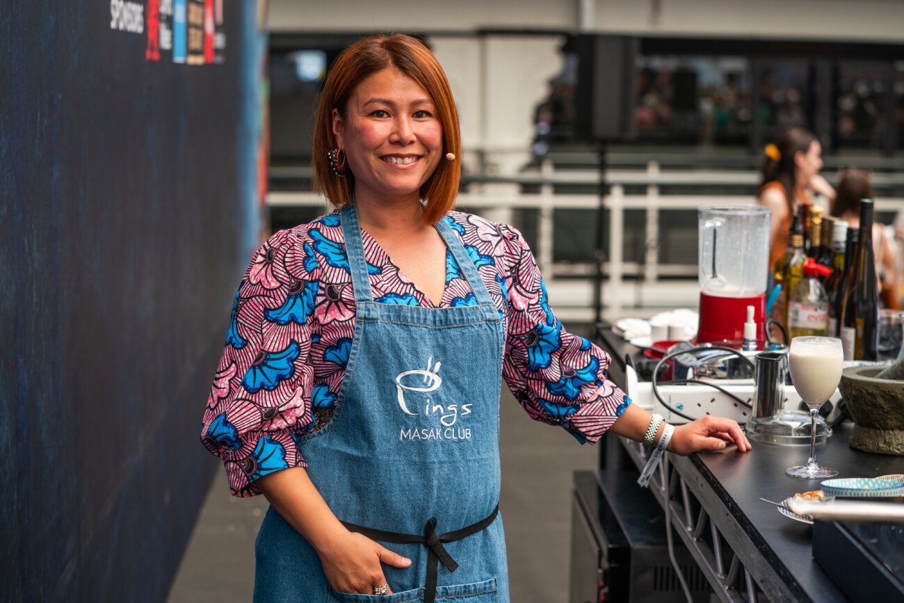 A smiling woman with shoulder-length hair wears a colorful blouse and a blue apron with "MASAK CLUB" on it. She stands by a kitchen counter with a blender and various ingredients in an open, bright setting. James Gifford-Mead Photography - Event Photographer London