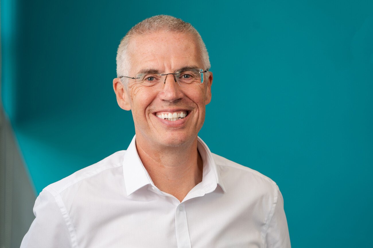 A smiling man with short gray hair and glasses, captured by a corporate headshot photographer, is wearing a white dress shirt in front of a teal background. James Gifford-Mead Photography - Event Photographer London