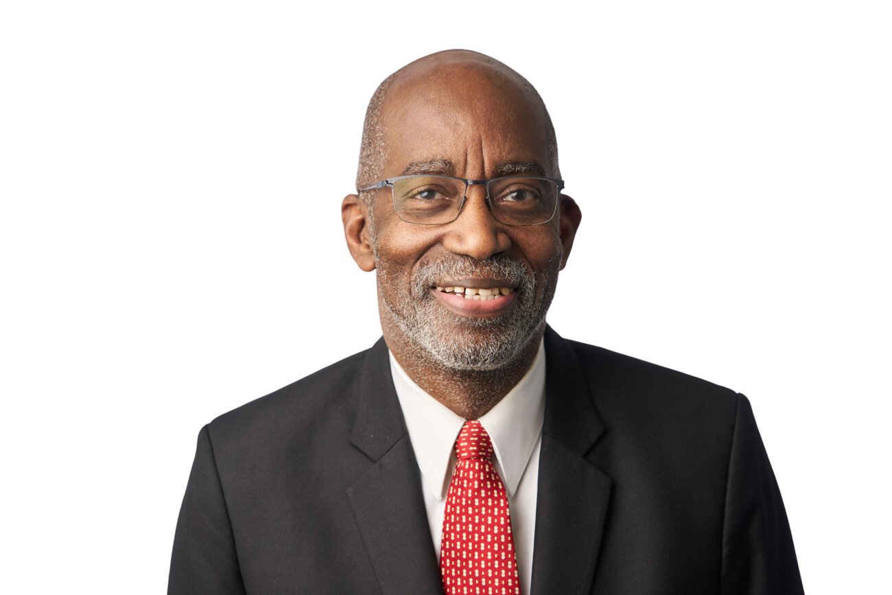 A corporate headshot photographer captures a smiling man with gray hair and a beard, wearing glasses, a black suit, white shirt, and red patterned tie against a pristine white background. James Gifford-Mead Photography - Event Photographer London