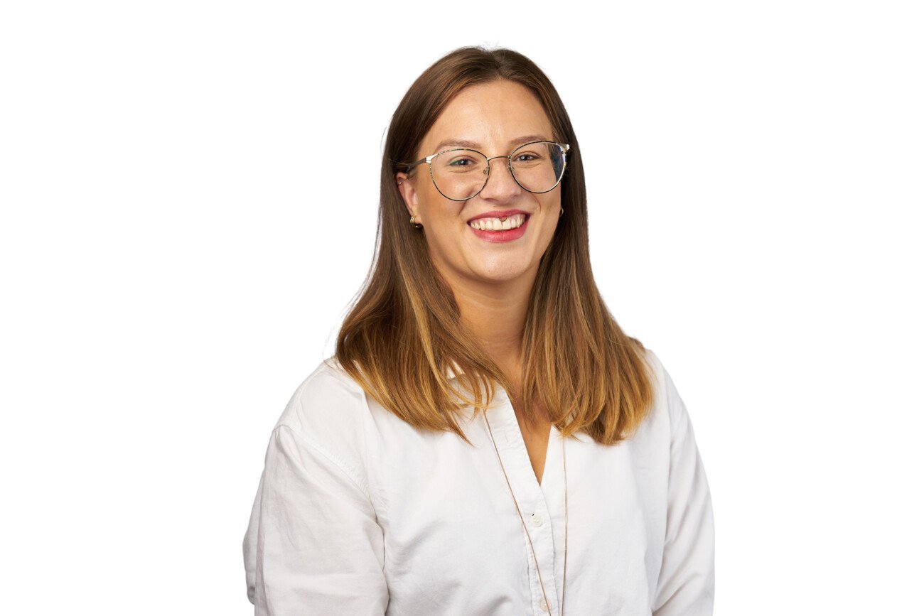 A person with glasses and long, straight hair smiles confidently at the camera, their white shirt complementing the crisp, plain background. This clean and professional image exudes the artistry of a skilled corporate headshot photographer. James Gifford-Mead Photography - Event Photographer London