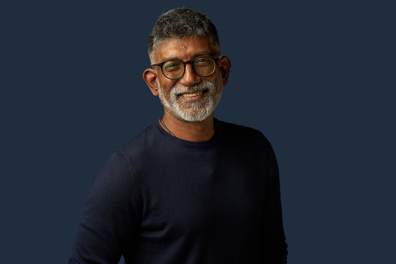 A smiling man with glasses, gray hair, and a beard stands against a dark background, wearing a dark long-sleeve shirt—a perfect capture by a corporate headshot photographer. James Gifford-Mead Photography - Event Photographer London