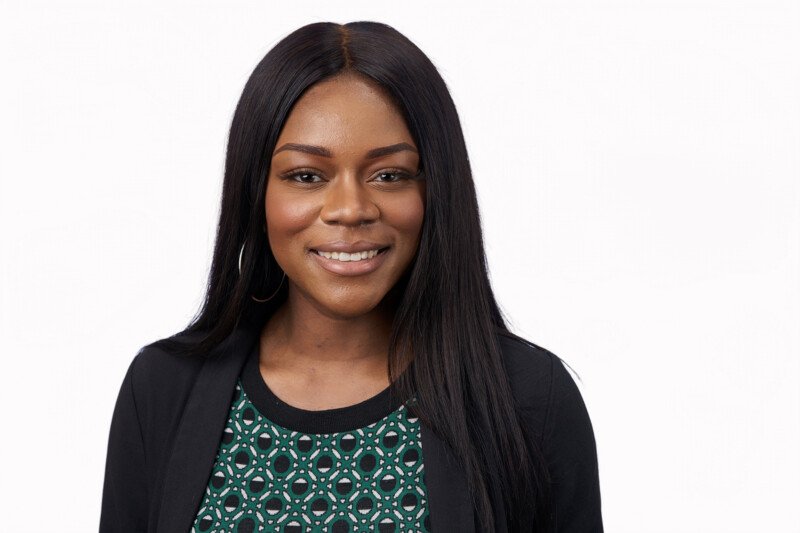 A smiling woman with long, straight black hair donning a black jacket and a green patterned top poses against a white background. Perfectly captured by a corporate headshot photographer, her expression exudes professionalism and warmth. James Gifford-Mead Photography - Event Photographer London