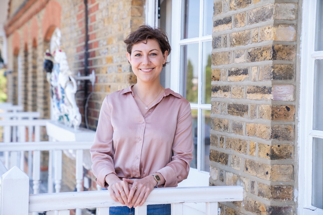 A person with short brown hair, in a light pink blouse and blue jeans, stands smiling on a balcony with brick walls and a white railing. Captured by a corporate headshot photographer, the scene features a decorative statue in the background. James Gifford-Mead Photography - Event Photographer London